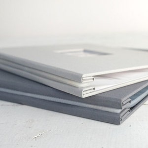 May include: Three photo albums stacked on top of each other. The top album is white with a rectangular window cut out of the cover. The middle album is white and the bottom album is gray.