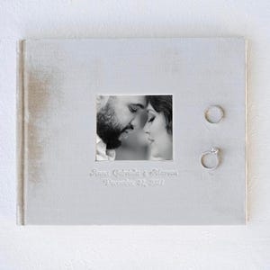 May include: A white wedding album with a black and white photo of a couple looking at each other. The album has the text "Anna Gabriella & Steven December 31, 2021" embossed on the front.