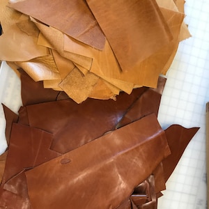 May include: A pile of brown leather scraps in various sizes and shapes. The leather is a light brown color and has a smooth texture.