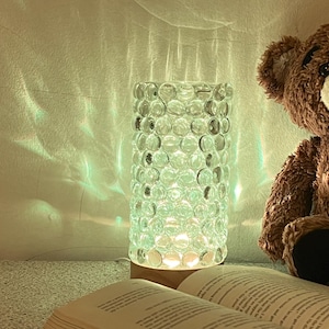 May include: A decorative lamp with a cylindrical glass shade filled with clear, round beads. The lamp sits on a wooden base and is next to a teddy bear and an open book. The lamp emits a soft, green glow.
