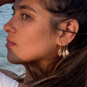 May include: Gold hoop earrings with seashell charms. The earrings are hanging from an ear with multiple piercings. The background is a blurred view of the ocean and sky.