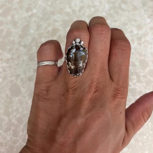 Peut inclure: Bague en argent ornée d'une grande pierre en forme de coquillage marron et blanc. La bague est surmontée d'une petite pierre transparente. Une autre bague en argent avec un motif floral est également visible.