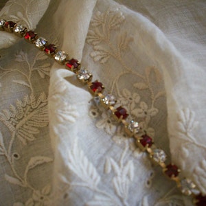 May include: A gold chain bracelet with alternating red and clear rhinestones. The bracelet is lying on a white fabric with an embroidered floral pattern.