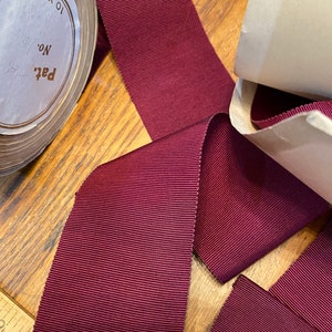Może przedstawiać: Rolka 10 jardów burgundowej wstążki grosgrain. Wstążka leży na drewnianej powierzchni i jest częściowo rozwinięta. Etykieta na rolce głosi "Pat's No. 8".