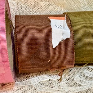 May include: Close-up of several rolls of ribbed ribbon in various colors. A pink ribbon roll is on the left, followed by a brown ribbon roll with a torn price tag, and a green ribbon roll. The ribbons are on a lace surface.