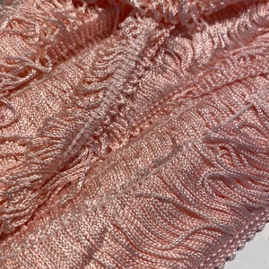 Puede incluir: Primer plano de un trozo de flecos rosa pálido. Los flecos están hechos de hilos suaves y sedosos, con un borde superior tejido. El color es un rosa claro delicado, y la textura parece suave y fluida. Este adorno es adecuado para varios proyectos de artesanía y costura.
