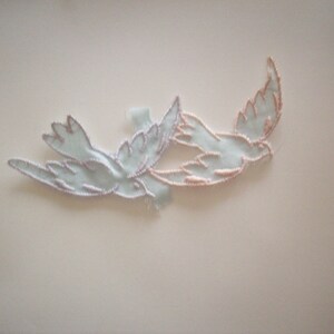 May include: Two embroidered birds in flight, one light blue and one peach, with a white background.