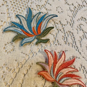 May include: Two embroidered flower patches, one in shades of blue, green, and orange, and the other in shades of pink, green, and orange.