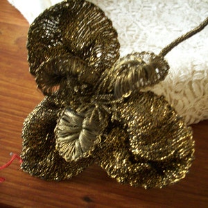 Puede incluir: Una flor de metal dorado, ornamental, con un diseño delicado e intrincado. La flor tiene varias capas de pétalos y un centro que se asemeja a un nudo.