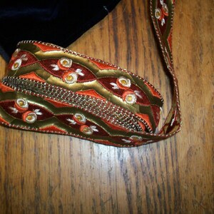 May include: A close-up of a gold, red, and white patterned ribbon with a beaded trim. The ribbon is folded and lying on a wooden surface.