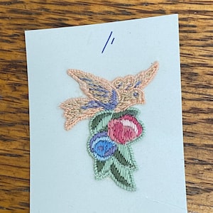 May include: Embroidered patch featuring a bird with peach and blue coloring perched above a cluster of pink, blue, and green berries. The patch is on a light blue background.