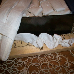 May include: Close-up of white elastic ribbon, partially coiled, with a portion of the ribbon extending across a wooden ruler. The ruler shows inch measurements. A dark box is in the background, filled with more ribbon.