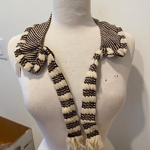 Puede incluir: Bufanda a rayas crema y marrón exhibida en un maniquí. La bufanda tiene un diseño tejido texturizado con un extremo con flecos. La bufanda es de color neutro y se puede usar con muchos atuendos.