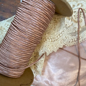 Puede incluir: Una bobina de cordón metálico de color oro rosa en una bobina de madera. El cordón está enrollado apretado y tiene un acabado brillante.