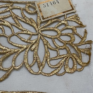 May include: A metallic gold lace trim featuring an intricate, openwork floral pattern with swirling, interconnected loops. A small, rectangular paper tag with a gold border is attached to the top, clearly displaying the black printed number 51464.
