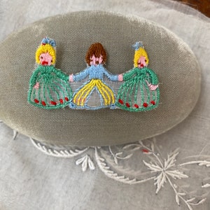 May include: Three embroidered figures of people in green, blue, and yellow dresses with red accents. The figures are sewn onto a light gray fabric.