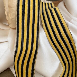 May include: Close-up of a decorative ribbon with vertical stripes. The ribbon features alternating black and gold stripes. The ribbon is made of a soft, velvety material. The ribbon is likely used for crafting, sewing, or embellishing various items.