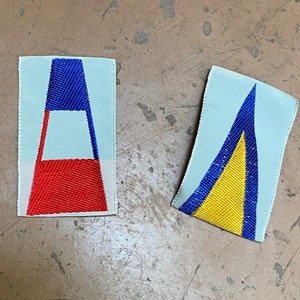 May include: Two rectangular fabric patches with geometric designs. One features a blue top, red bottom, and white center. The other has a blue and yellow design. Both are on a light gray background.
