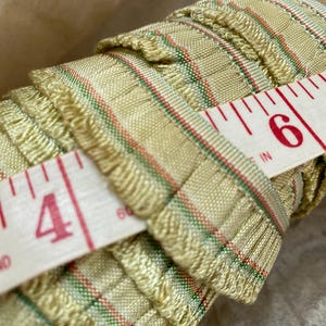 May include: A roll of beige trim with green and red stripes. The trim has a fringed edge. A measuring tape is wrapped around the trim, showing the numbers 4 and 6 inches.