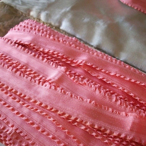 Op de afbeelding: Close-up van een rol roze lint met een getextureerde, geschulpte rand. Het lint is gelaagd en toont de gedetailleerde weefsel en kleurvariaties. Andere stofresten zijn zichtbaar op de achtergrond.