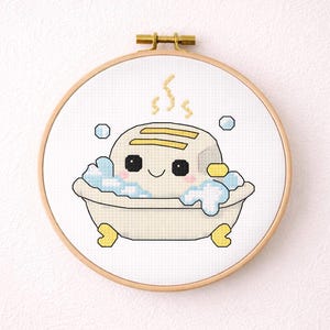 May include: Cross-stitch embroidery of a smiling toaster in a bathtub with bubbles. The toaster has yellow accents and is surrounded by blue bubbles and suds. The embroidery is set in a round wooden hoop.
