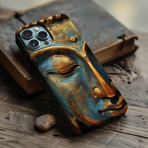 Puede incluir: Una funda de teléfono con un diseño detallado de la cara de Buda. La funda tiene un aspecto texturizado con tonos dorados y azules, y está sobre una superficie de madera junto a un libro.