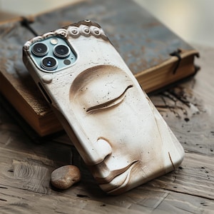 Puede incluir: Una funda de teléfono blanca y beige con un relieve detallado de un rostro sereno de Buda. La funda tiene un aspecto texturizado y envejecido, con las lentes de la cámara del teléfono visibles. La funda descansa sobre una superficie de madera, junto a una pequeña piedra y un libro antiguo.