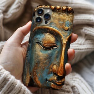 May include: A detailed phone case featuring a serene Buddha face in shades of teal and gold. The case has a textured, three-dimensional design with intricate details. The phone case is held in a person's hand, with a soft, neutral-colored background.
