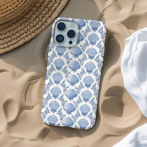 May include: A white and blue phone case featuring a seashell pattern. The case has a repeating design of blue seashells and leafy branches on a white background. The phone case is on a sandy surface.