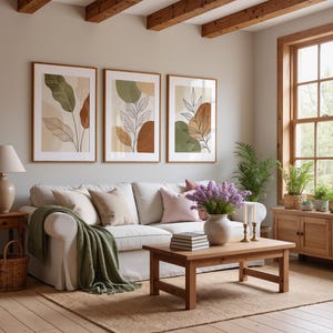 Puede incluir: Tres impresiones art&iacute;sticas bot&aacute;nicas enmarcadas con dise&ntilde;os abstractos de hojas en tonos verdes, marrones y beige cuelgan sobre un sof&aacute; blanco. Una mesa de centro de madera, un jarr&oacute;n con flores moradas y una manta verde completan el acogedor sal&oacute;n.