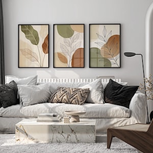 Puede incluir: Tres impresiones art&iacute;sticas bot&aacute;nicas enmarcadas con dise&ntilde;os abstractos de hojas en tonos verdes, marrones y beige cuelgan sobre un sof&aacute; blanco. El sal&oacute;n cuenta con una mesa de centro de m&aacute;rmol, cojines decorativos y una silla de madera.