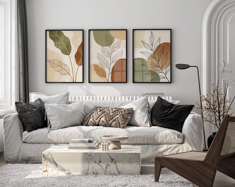Puede incluir: Tres impresiones art&iacute;sticas bot&aacute;nicas enmarcadas con dise&ntilde;os abstractos de hojas en tonos verdes, marrones y beige cuelgan sobre un sof&aacute; blanco. El sal&oacute;n cuenta con una mesa de centro de m&aacute;rmol, cojines decorativos y una silla de madera.