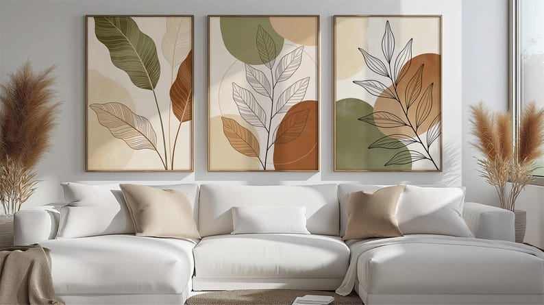 Puede incluir: Tres impresiones art&iacute;sticas bot&aacute;nicas enmarcadas con dise&ntilde;os abstractos de hojas en tonos verdes, marrones y beige. Las impresiones se exhiben sobre un sof&aacute; seccional blanco con cojines de colores neutros. Hierba de la pampa decorativa en jarrones a ambos lados.