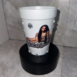 May include: White foam cup decorated with a photo of a woman, rhinestones, and pearls. The cup has the text "Chapter 2" in a cursive font. The cup is on a black stand.