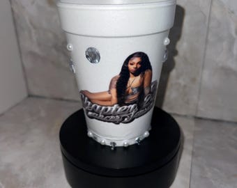 Può includere: Tazza di schiuma bianca decorata con una foto di donna, strass e perle. La tazza ha il testo "Chapter 2" in corsivo. La tazza è su un supporto nero.