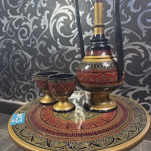 May include: A decorative, hand-painted wooden table with a matching jug and two small cups. The table and serving pieces are black, gold, and red with intricate patterns. The jug has a long handle and a slender neck.