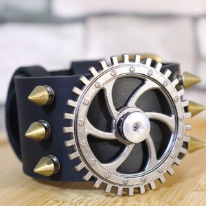 May include: Black leather cuff bracelet featuring a silver gear design and gold-colored cone studs. The steampunk-inspired accessory has a buckle closure and is set against a wooden surface.