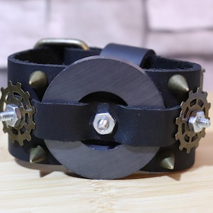 May include: Black leather steampunk-style bracelet with a central dark gray ring and brass gear accents. The bracelet features brass-colored cone studs and a metal buckle. The design incorporates industrial and mechanical elements.