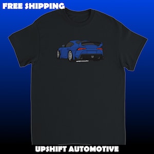 Puede incluir: Una camiseta negra de manga corta con una ilustración gráfica de un coche deportivo azul visto desde un ángulo trasero de tres cuartos. El diseño del coche incluye un alerón trasero y el texto "upshift automotive" en letras blancas.