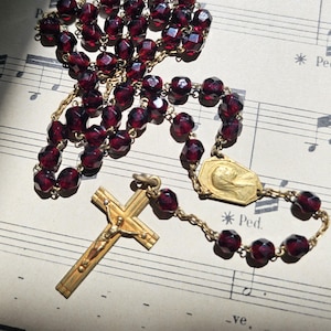 May include: A rosary featuring deep red, faceted beads and gold-coloured metal components. The cross and connecting elements are gold. The rosary is displayed on a sheet of vintage sheet music with text.