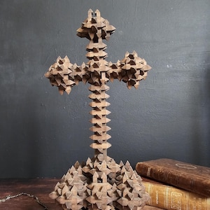 Könnte beinhalten: Eine detaillierte Holzkreuzskulptur mit geometrischem Design. Das Kreuz ist braun und steht aufrecht, mit komplizierten, stacheligen Details. Ein Rosenkranz und antike Bücher befinden sich im Hintergrund, was auf ein religiöses oder historisches Thema hindeutet.