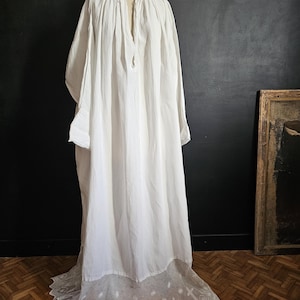 May include: A long, white, vintage-style nightgown with long sleeves and a high neckline. The gown features a delicate, embroidered lace hemline with a cross and star pattern. The garment is displayed on a mannequin against a dark background.