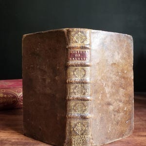 May include: A vintage, brown leather-bound book with gold accents and the title "HISTOIRE DE SALUSTE" on the spine.
