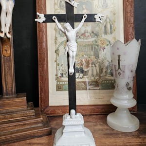 May include: A white porcelain crucifix with a black cross and a white figure of Jesus. The cross has decorative white accents and the inscription "INRI". The crucifix is on a white base with ornate carvings. A framed religious print is in the background.