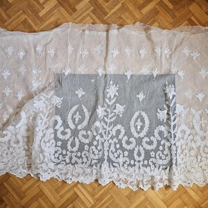 May include: A sheer off-white lace fabric featuring an intricate white floral embroidery pattern. The design includes scattered small flower motifs across the upper section and a dense, elaborate botanical border along the bottom edge.