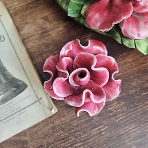 May include: A burgundy ceramic flower with a glossy finish. The flower has multiple petals and is surrounded by green leaves. A vintage book is visible in the background. The flower is likely a decorative item.