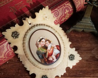May include: A white, ornate frame with a carved leaf design surrounds a miniature painting of the Virgin Mary holding a baby Jesus. The painting is set against a red and gold background.