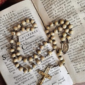 May include: An antique rosary featuring cream-coloured beads and a gold crucifix rests on the open pages of an old book. The book's aged pages are filled with faded text, enhancing the vintage feel.