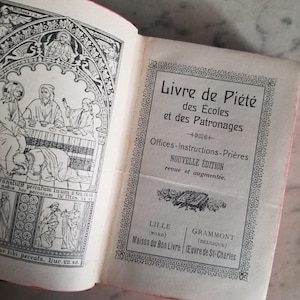 Può includere: Libro aperto con illustrazioni e testo in bianco e nero. Il titolo del libro è "Livre de Piété des Écoles et des Patronages". Le pagine hanno un bordo rosa. Il libro è aperto su una pagina con disegni dettagliati e testo.