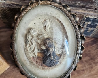 May include: Vintage oval religious medallion with a detailed relief scene under a glass cover. The medallion has a decorative, scalloped edge and a tarnished brass finish. The scene depicts figures in prayer, with text at the bottom.